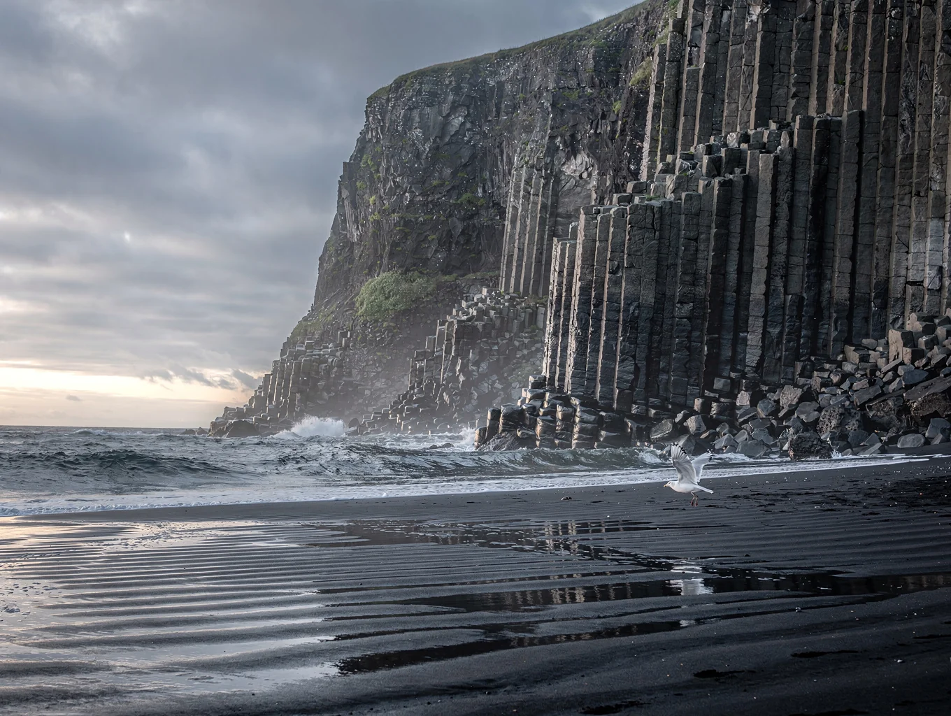 Long exposure ocean waves washing over black volcanic sand and geometric basalt column cliffs